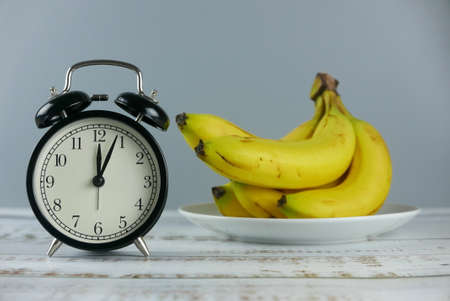 Selective focus bunch of bananas and alarm table clock show 4 past 12 o'clock on wooden background.の写真素材