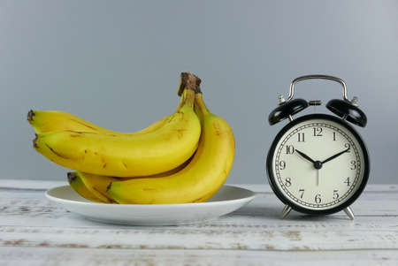 Selective focus bunch of bananas and alarm table clock show 10 past 10 o'clock on wooden background.