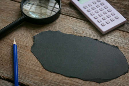 Pencil,magnifying glass and calculator with empty piece of black paper on wooden background. Copy...