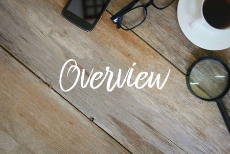 Top view of magnifying glass,a cup of coffee,glasses and mobile phone on wooden background written with overview.の写真素材
