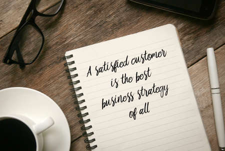 Top view of a cup of coffee.glasses,mobile phone,pen and notebook written with quote A satisfied customer is the best business strategy of all.の写真素材