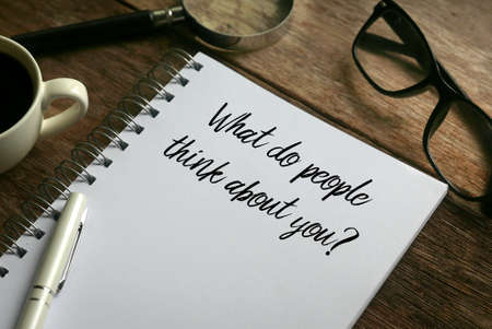 Selective focus of coffee,magnifying glass,glasses,pen and notebook written with question of What do people think about you? on wooden background.の写真素材