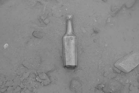 Selective focus of an old glass bottle covered with mud and water by the beach. Photo in black and...