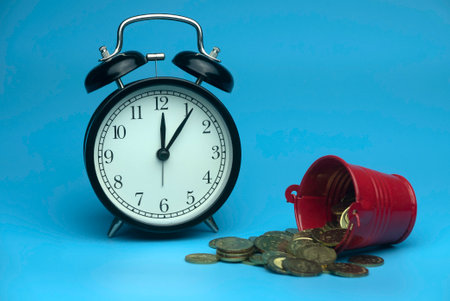 Selective focus of table clock and a red bucket with a gold coins isolated on a blue background. Business and finance concept.の写真素材
