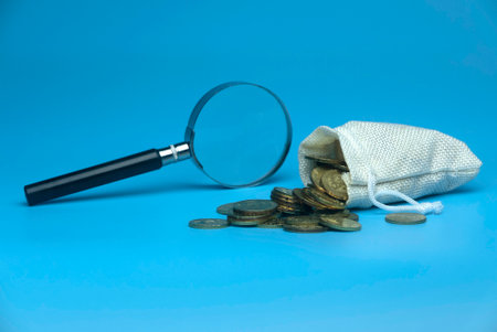 Selective focus of magnifying glass and burlap sack full with gold coins on blue background. Business and finance concept.の写真素材