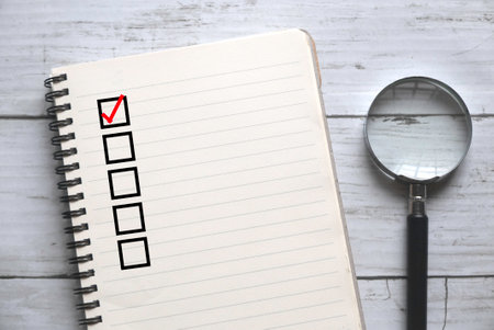 Top view of a magnifying glass and book with blank checklist on a white wooden background.の写真素材