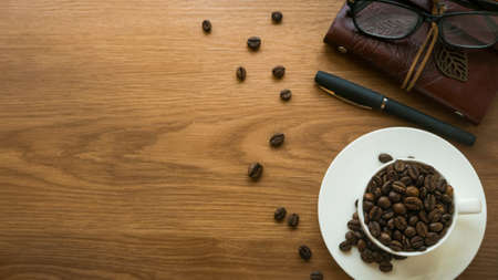 Top view of a white cup with a coffee bean, glasses, notebook and pen on wooden background with copy...