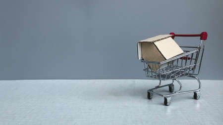 Real estate concept. A shopping cart with a house model on it on grey background with copy space.