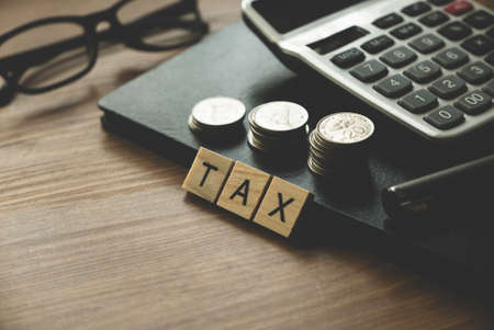 Tax concept. Selective focus of wooden alphabet with stack of coins pen, calculator,glasses and notebook on wooden background.の写真素材