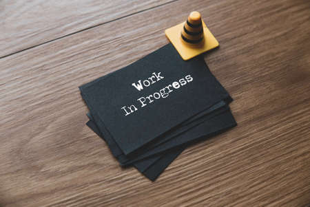 Selective focus of yellow safety cone and a stack of black paper written with Work In Progress on wooden background.の写真素材