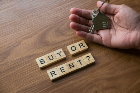Buy or rent concept. Selective focus of wooden alphabet and a hand holding a key with house keychain on wooden background.の写真素材