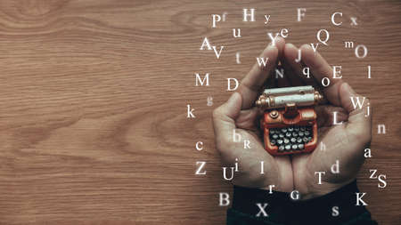 A hand holding toy typewriter with alphabet flying around on wooden table with copy space.の写真素材