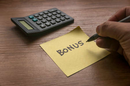 Selective focus of hand writing a word Bonus in yellow paper with calculator on wooden background.