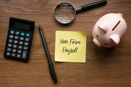 Top view of calculator,magnifying glass, piggy bank, pen and yellow memo paper written with Non Farm Payroll on wooden background.の写真素材