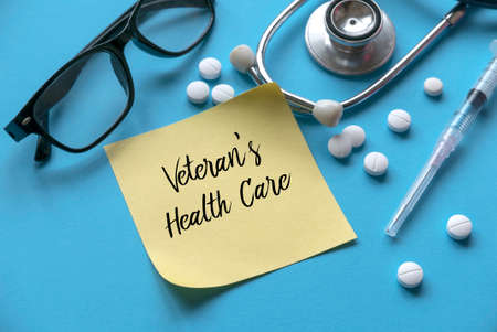 A glasses, pills, syringe, stethoscope and yellow memo sticky note written with Veteran's Health Care on blue background.の写真素材