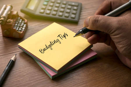 A hand writing Budgeting Tips on a yellow memo note with calculator and miniature typewriter.
