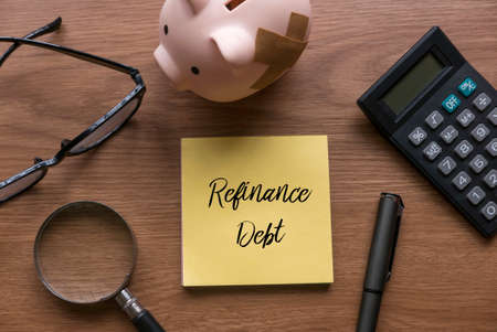 Top view of glasses, magnifying glasses, pen, piggy bank, calculator and memo note written with Refinance Debt on wooden background.の写真素材