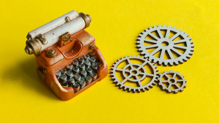 A miniature vintage typewriter next to wooden gears, symbolizing the integration of old and new technology, mechanical writing process, and industrial innovation on a bright yellow background.の写真素材
