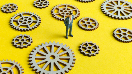 Miniature businessman standing among scattered wooden gears on a bright yellow background,...