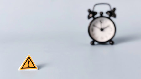 Yellow Warning Sign and Blurred Alarm Clock on Blue Background Highlighting Time Constraints, Danger, and Urgent Deadlinesの写真素材