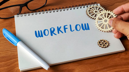 Hand adjusting wooden gears next to the word Workflow written in a notebook with a marker and glasses on a desk.の写真素材