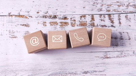 Wooden Blocks with Contact Icons (Email, Phone, Chat) Lined Up on a White Wooden Desk, Symbolizing Communication Methods and Customer Serviceの写真素材