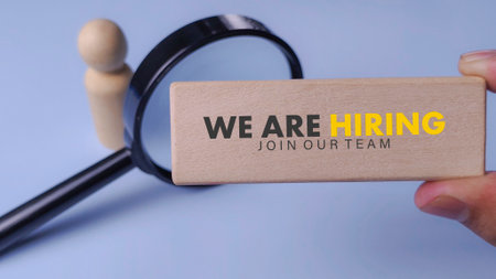 We Are Hiring: Wooden Block Held Over Magnifying Glass and Wooden Figure for Recruitment and Job Searchの写真素材