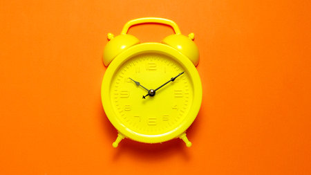 Vibrant yellow alarm clock centered on a bright orange background for high contrast conceptsの写真素材
