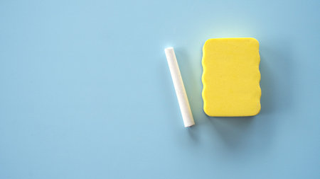 Top-down view of a white chalk stick and a yellow foam sponge placed on a solid light blue surface.
