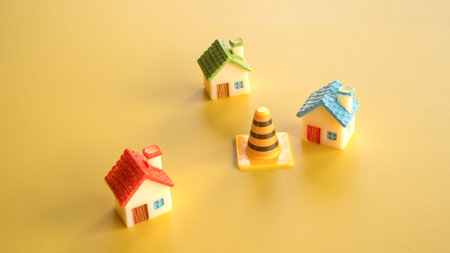 Three miniature colorful toy houses arranged around a single orange and black striped traffic safety cone on a smooth yellow surface.