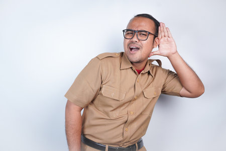 a man Asian civil servants or state civil servants is with brown uniform. gesture trying to hear you overhear listening intently. On white background. Indonesia PNS, ASNの写真素材