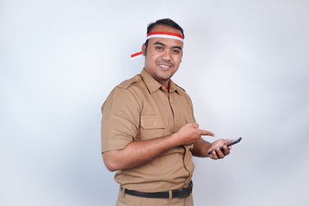 a man Asian civil servants or state civil servants is with brown uniform and red and white Headband gesture headband, holding his phone, and pointing copy space beside his. On white background. Indonesia PNS, ASNの写真素材