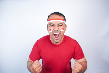 A young Asian man with a happy successful expression, raised a fist wearing red shirt and flag headband isolated by white background. Indonesia's independence day concept.の写真素材