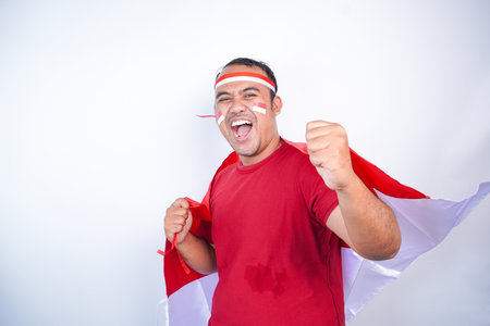 Asian young man using a flag clenched both first to celebrate Indonesia independence day on August 17, looking at camera with confident and excited expression isolated on white backgroundの写真素材