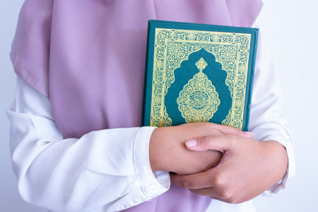 A Muslim woman wearing a white Muslim dress holds an Al-Qur'an Muslim holy book on her chest. the cover of the book is written as "Al-Qur'anul Karim" in Arabic calligraphy. isolated white backgroundの写真素材