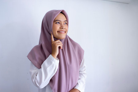 young asian muslim woman wearing a purple hijab, scarf smiling and pointing to her cheek think. Concept of warmth and friendliness, as the woman is comfortable and happy in her surroundings.の写真素材
