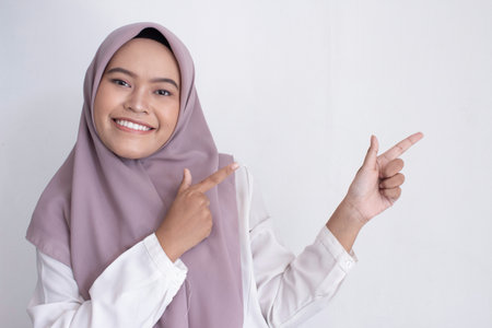 young asian muslim woman wearing a purple hijab, scarf smiling is pointing to the right. She has a big smile on her faceの写真素材