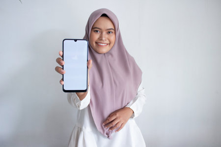 young muslim asian woman wearing a purple hijab. a cell phone and pointing to the screen. She is...