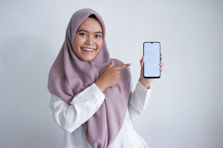 young muslim asian woman wearing a purple hijab. a cell phone and pointing to the screen. She is...