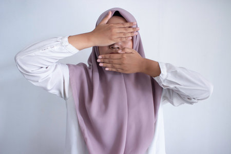 young asian muslim woman wearing a purple hijab purple scarf is covering her face with her hands. Concept of mystery and intrigue, as the woman's actions suggest that she is hiding something or trying to conceal her emotionsの写真素材