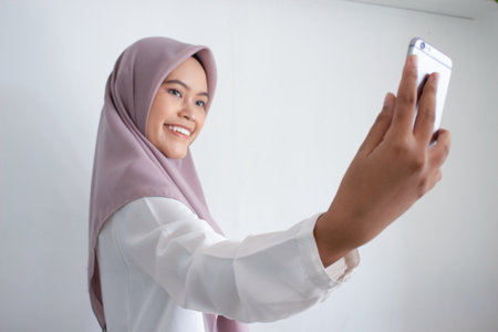 young muslim asian woman wearing a purple hijab head scarf is taking a selfie with her phone. She is smiling and she is happyの写真素材
