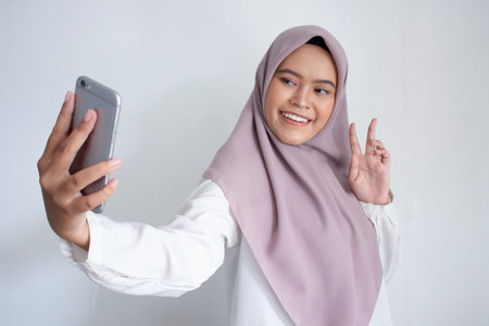 young muslim asian woman wearing a purple hijab head scarf is taking a selfie with her phone. She is smiling and she is happyの写真素材