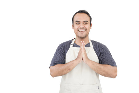 Young smiling Asian man as grocery store staff standing at cashier counter with arms namaste pose and full of confidence in front of display shelvesの写真素材