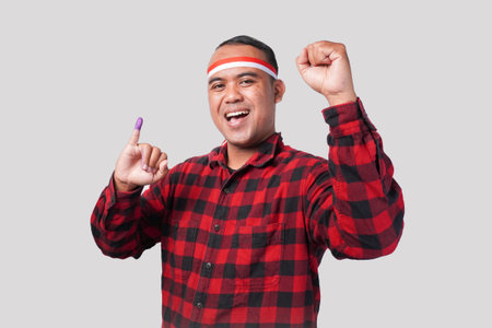 Energetic Indonesian man in flannel shirt outfit, flag headband,, proudly showing little finger dipped in purple ink after voting for electionの写真素材