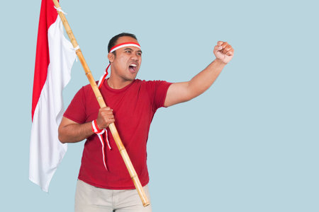 young Energetic Indonesian man is holding a flag while raising fist gesture and shouting loud, Indonesia Independence Day concept.の写真素材