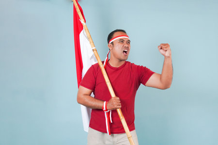 young Energetic Indonesian man is holding a flag while raising fist gesture and shouting loud,...