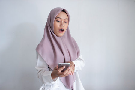 young muslim asian woman wearing a purple hijab, head scarf is holding a cell phone. She is looking...