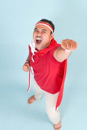Happy young Indonesian man wearing country flag on her back and raising hand superhero fly gesture. Indonesia Independence Day concept.の写真素材
