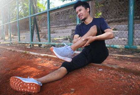 A male runner, sportsman sits on the roadside in sportswear and holds his leg, cramps, feels severe...