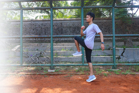 A man is doing a leg exercise in front of a fence. The fence is green and has a stone wall behind...